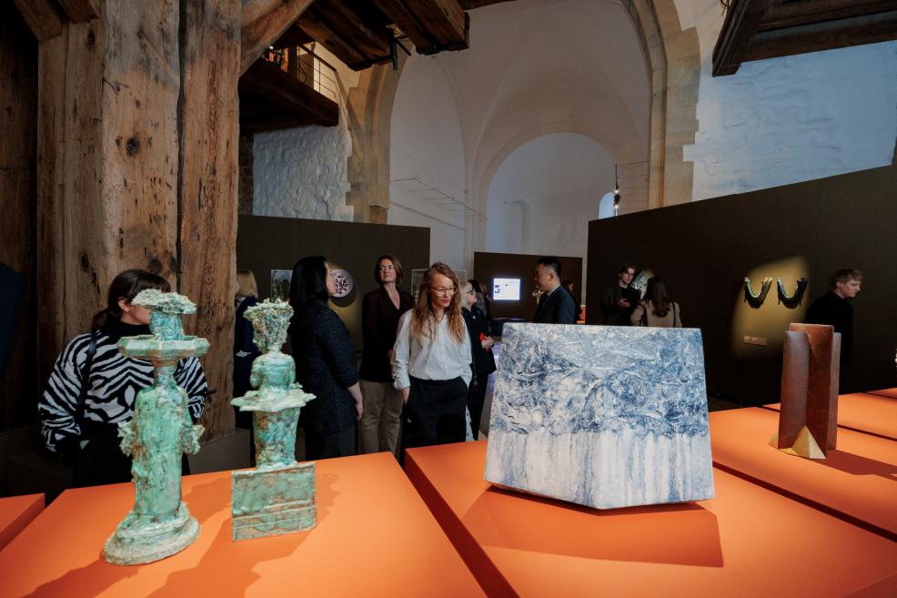 Exhibition hall with visitors, featuring abstract sculptures on orange pedestals and large wooden beam structures.