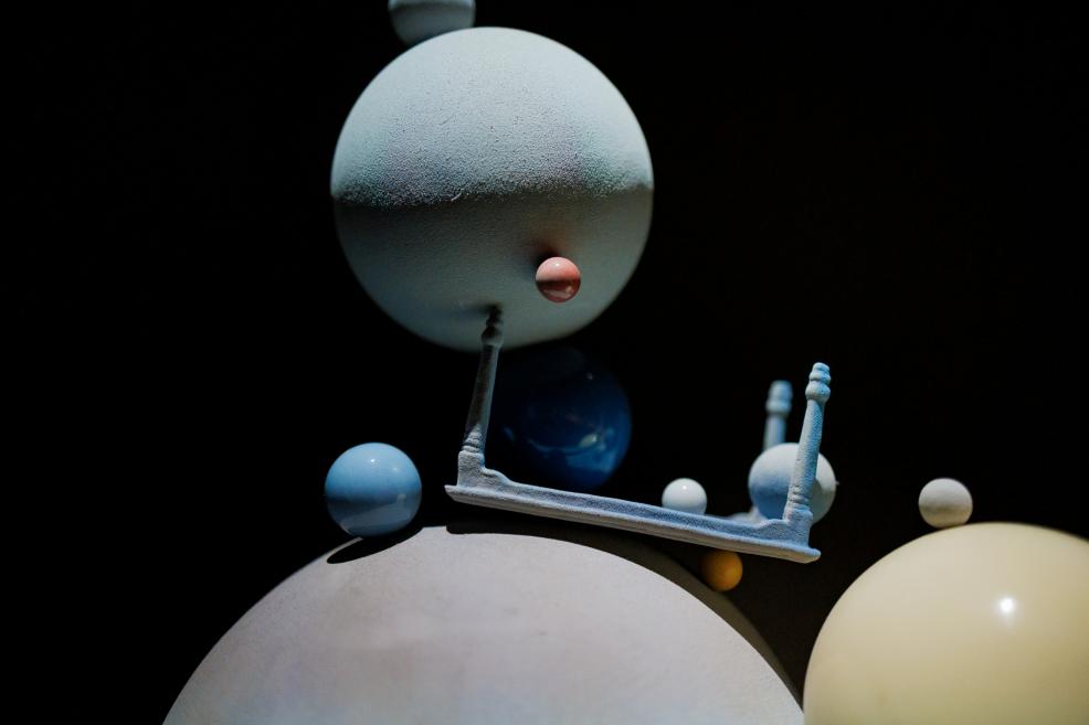 An abstract installation against a dark background features large and small spheres in light blue, white and yellow tones, along with a white horizontal structure.