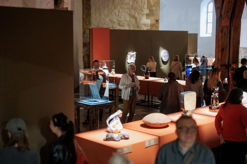 Visitors in a museum exhibition hall stand among orange pedestals with ceramic and glass artworks in display cases.