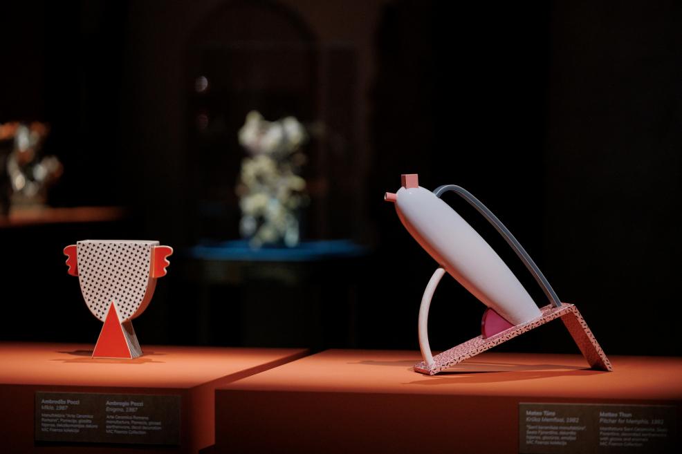 Two abstract ceramic objects with geometric forms are displayed on an orange platform in a museum exhibition against a dark background.