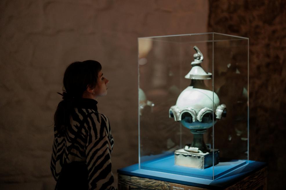 A dark-haired woman in a zebra-patterned sweater looks at a small sculptural object displayed in a glass case on a blue base.