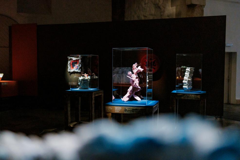 Several small sculptures and objects are displayed in glass cases on blue plinths in a dark exhibition hall.