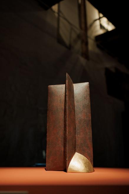 Abstract bronze sculpture with vertical facets on an orange surface in a dark room.