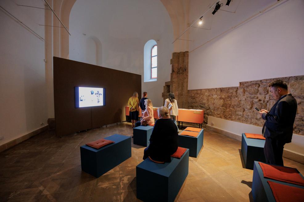 Museum exhibition room with visitors looking at a wall-mounted screen and blue seating blocks with orange cushions.