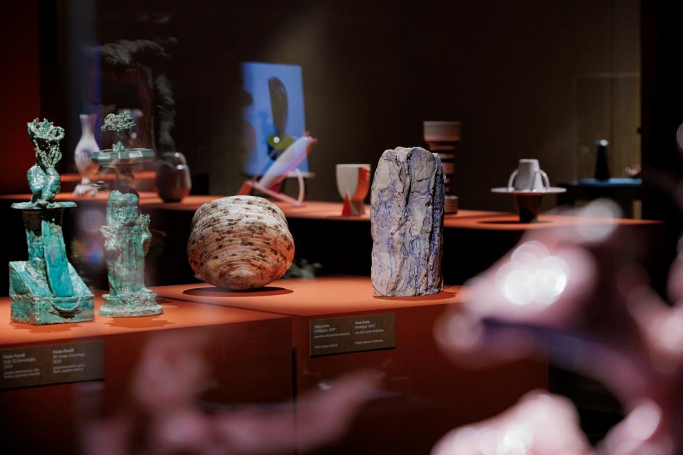 Several contemporary ceramic and sculptural objects in different shapes and colors are arranged in a museum display case on orange pedestals.