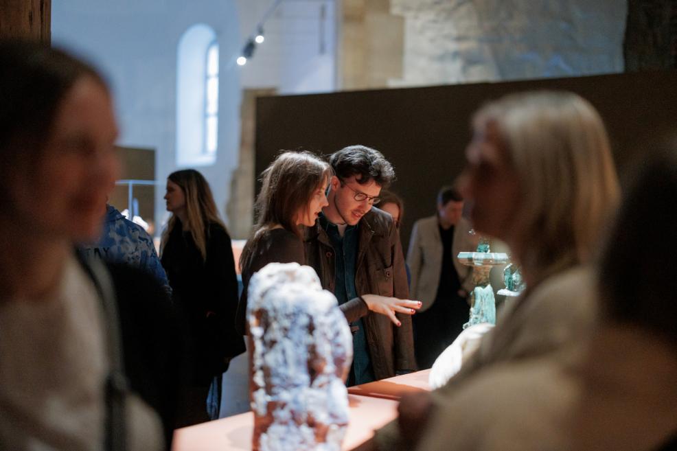 In a museum gallery, several visitors view the exhibition, with a man wearing glasses and a woman standing by sculptures on a lit table.