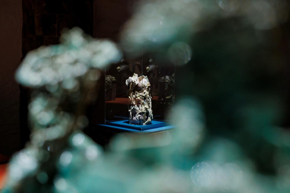 An abstract sculpture stands on a blue pedestal inside a museum display case, partially obscured in the foreground by blurred greenish light reflections.