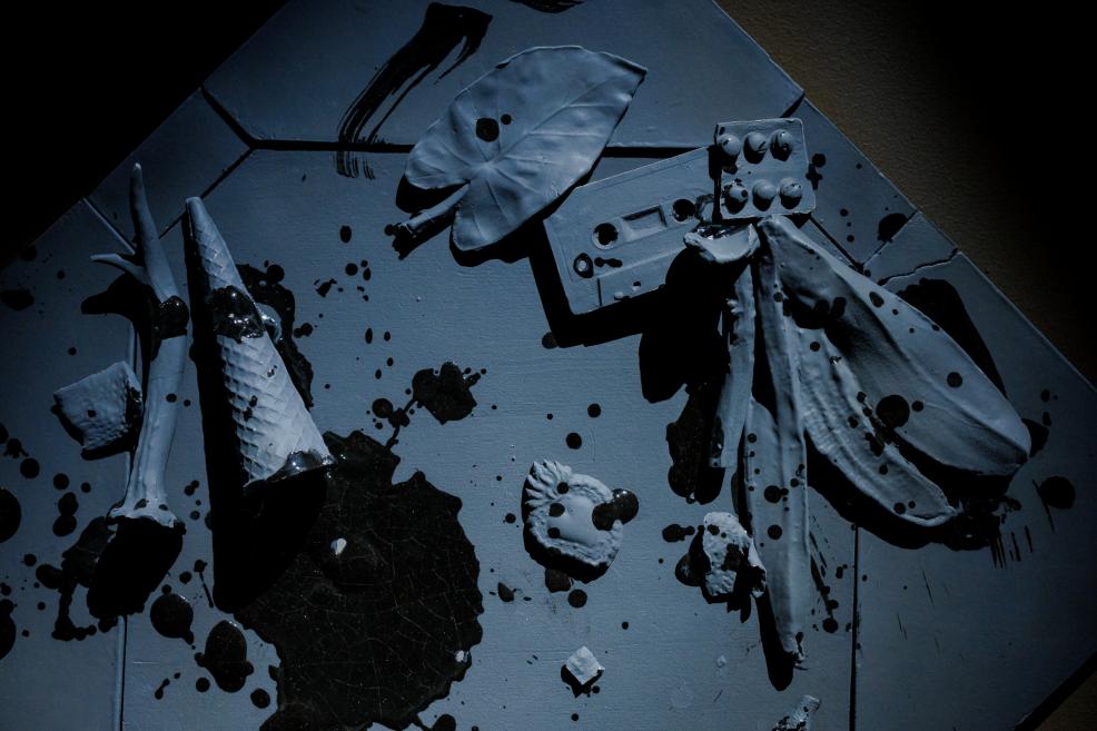 Abstrakta zila reliefa kompozīcija ar piestiprinātiem baltiem veidojumiem un melniem krāsas šļakstiem uz daudzstūra formas pamatnes.