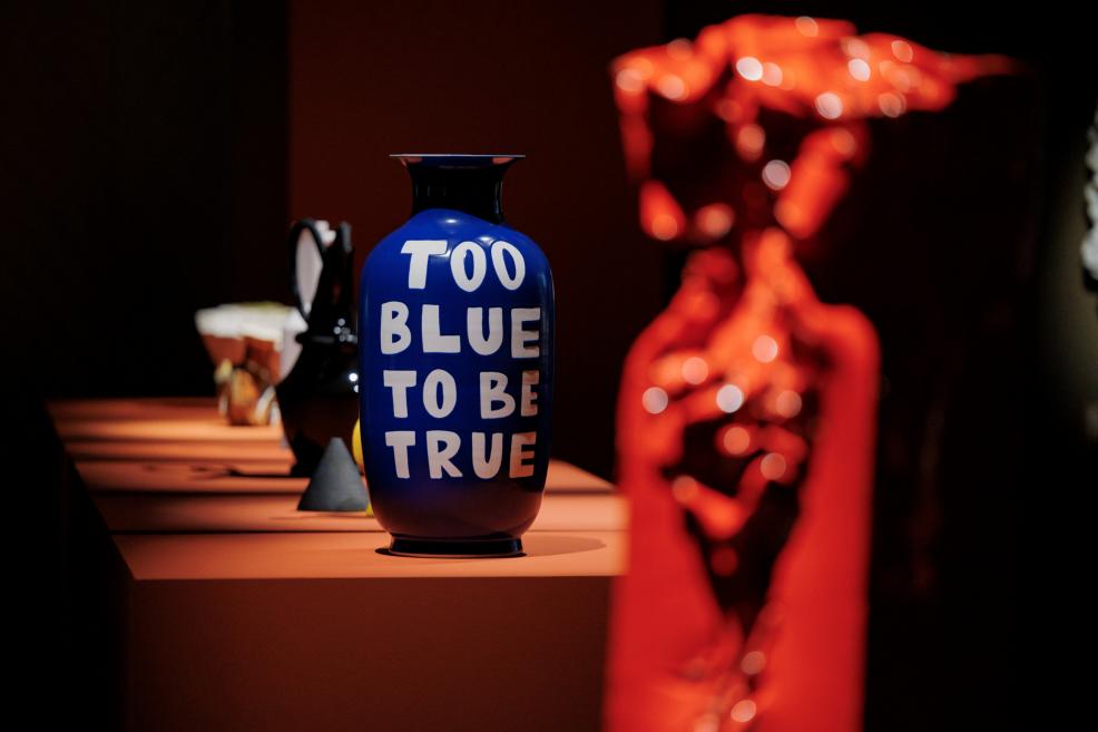 Zilā vāzē ar uzrakstu TOO BLUE TO BE TRUE uz tumša pjedestāla izstādē redzami vairāki keramikas objekti, tostarp sarkani, izplūduši priekšplānā.
