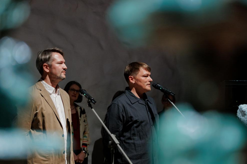 Divi vīrieši stāv pie mikrofoniem uz skatuves ar vēl vairākiem cilvēkiem fonā, priekšplānā redzami izplūduši zili gaismas plankumi.