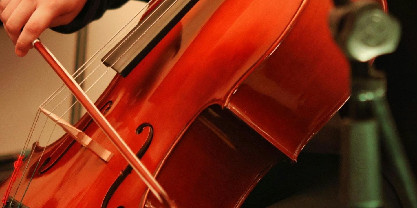 Tuvplānā redzams cilvēks, kas ar lociņu spēlē čellu, bet priekšplānā redzama mikrofona statīva daļa.