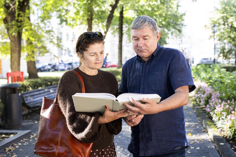 Divi cilvēki ārā parkā kopīgi skatās atvērtu grāmatu, vīrietim turot to rokās.