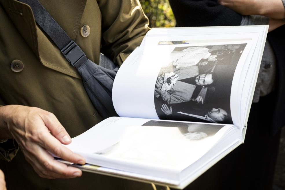 Persona ar melnu plecu somu rokās tur lielu atvērtu fotogrāmatu ar melnbaltām cilvēku fotogrāfijām.