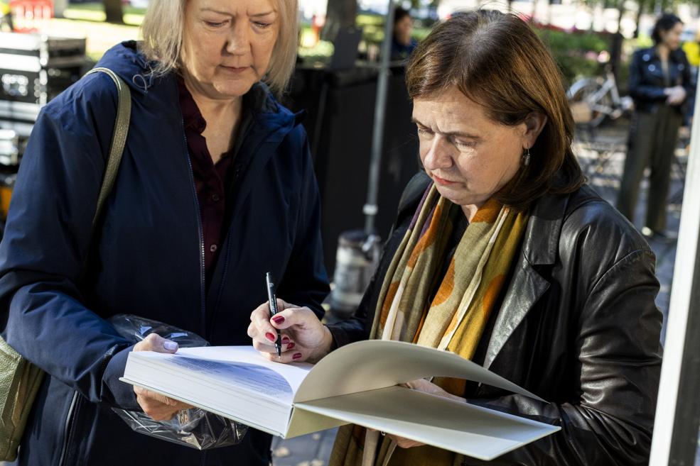 Divas sievietes ārā apskata un paraksta lielu grāmatu, viena to tur rokās, kamēr otra raksta ar pildspalvu.