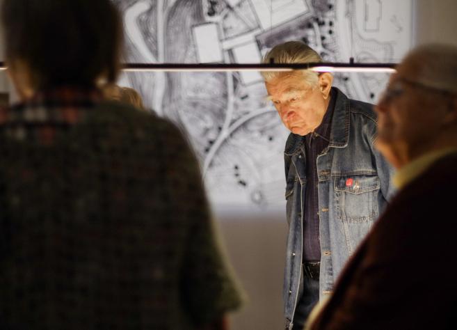 Apmeklētāji muzeja ekspozīcijā aplūko izgaismotu karti vai plānu uz galda.