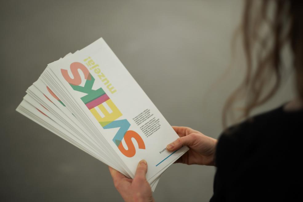 Kaudzītē sakārtotas krāsainas muzeja izglītojošās kartītes ar uzrakstu “Sveiks muzejā!”.