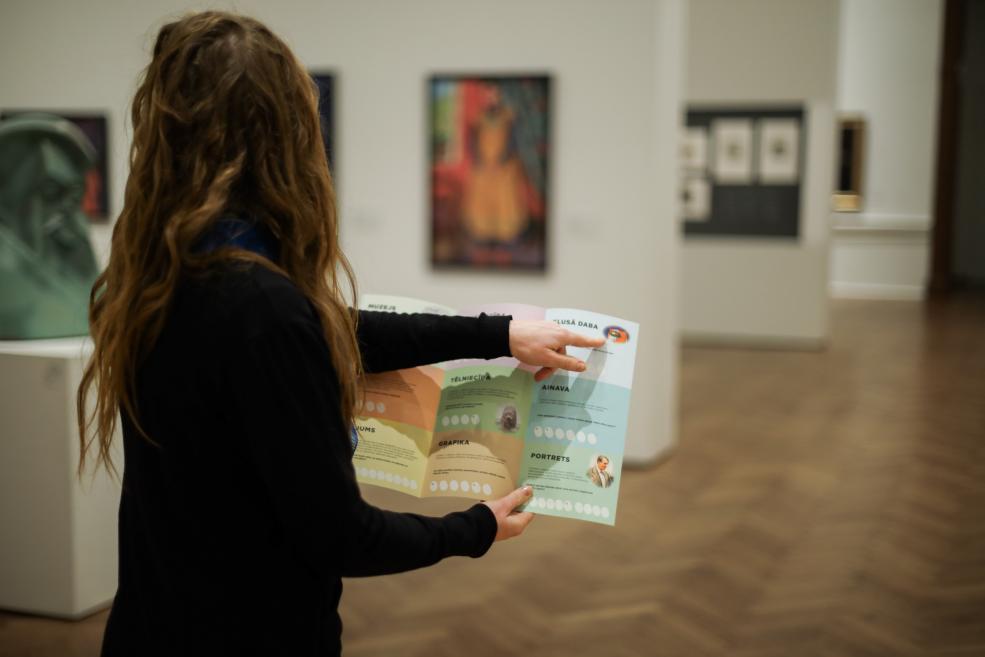 Apmeklētāja muzejā tur atvērtu izglītojošu karti un norāda uz uzdevumu sadaļu, fonā redzami mākslas darbi ekspozīcijā.