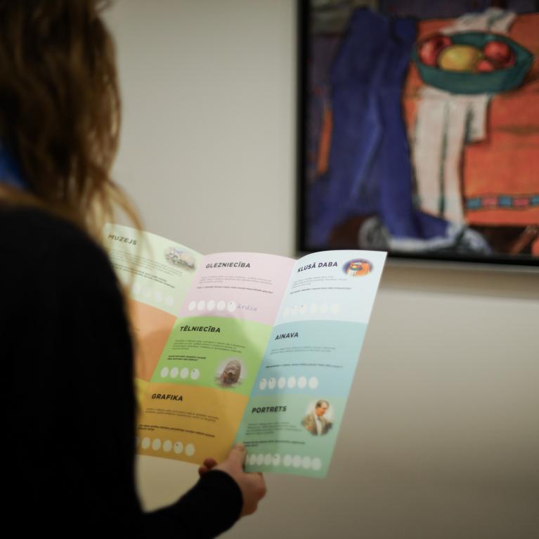 Apmeklētāja muzejā skatās izglītojošu uzdevumu karti, fonā redzama glezna ar kluso dabu ekspozīcijā.