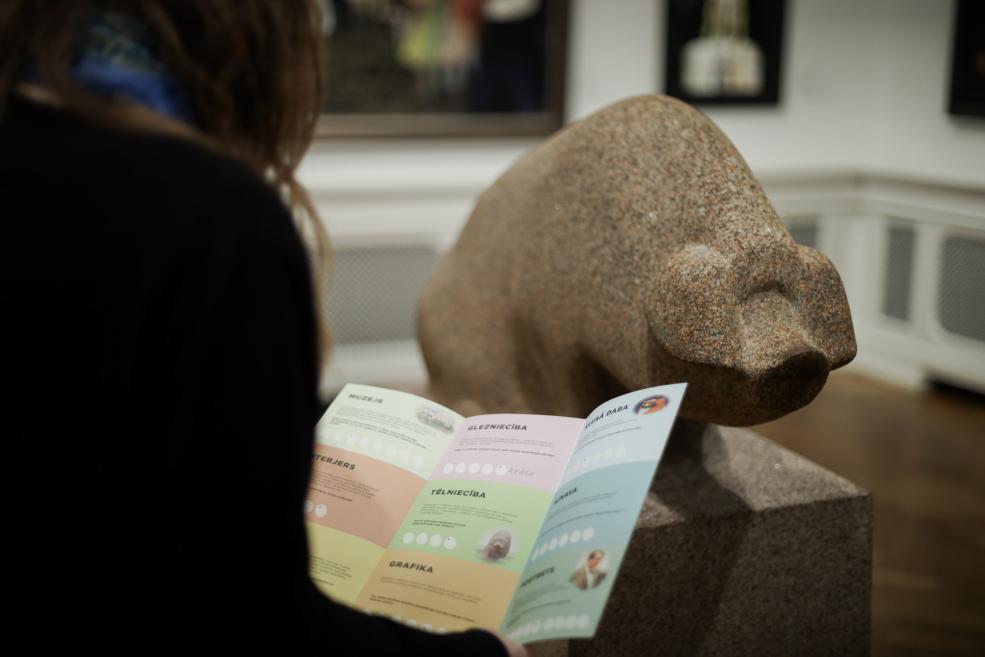 Apmeklētāja muzejā skatās izglītojošu uzdevumu karti, fonā redzama glezna ar kluso dabu ekspozīcijā.