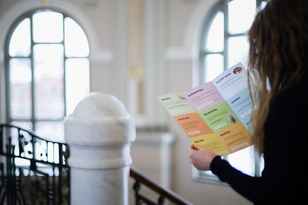 Apmeklētāja muzeja kāpņu telpā lasa izglītojošu uzdevumu karti, fonā redzami augsti logi un vēsturiska interjera detaļas.