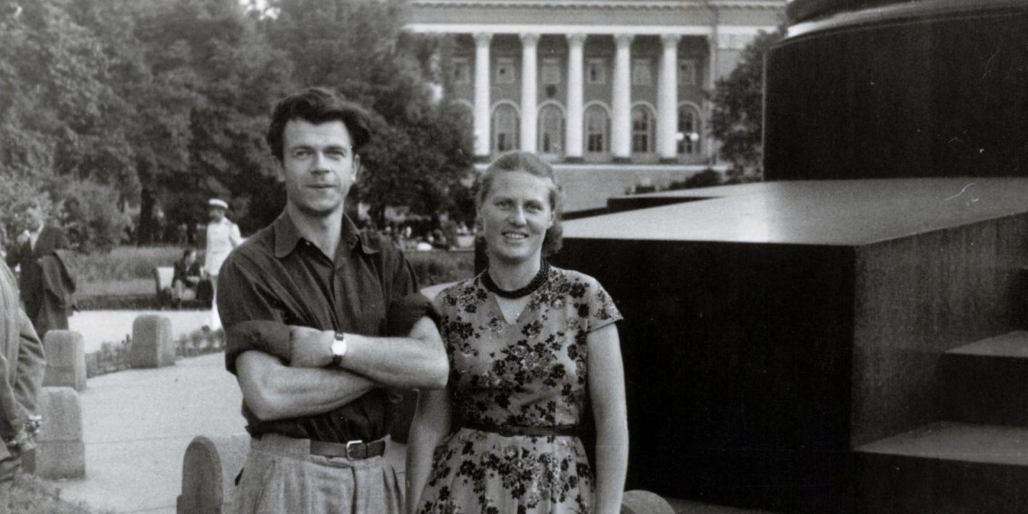 Melnbalta fotogrāfija ar jaunu vīrieti un sievieti, kas stāv parkā pilsētvidē; fonā redzama monumentāla ēka ar kolonnām.