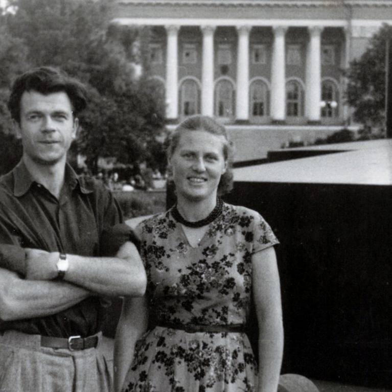 Melnbalta fotogrāfija ar jaunu vīrieti un sievieti, kas stāv parkā pilsētvidē; fonā redzama monumentāla ēka ar kolonnām.