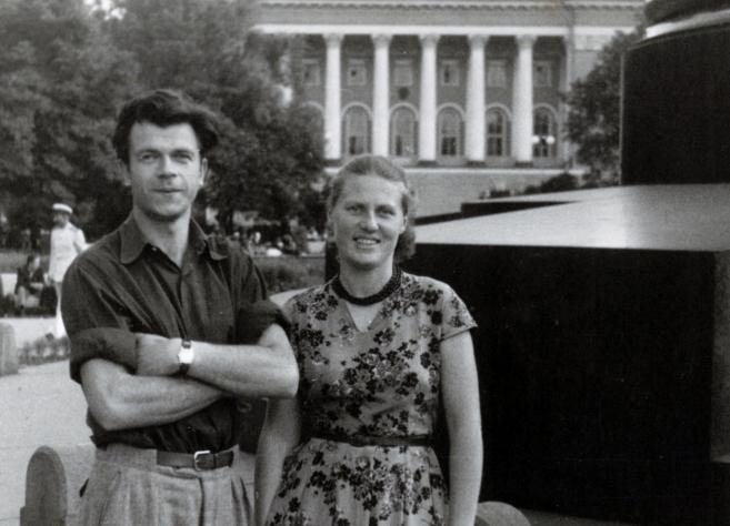 Melnbalta fotogrāfija ar jaunu vīrieti un sievieti, kas stāv parkā pilsētvidē; fonā redzama monumentāla ēka ar kolonnām.