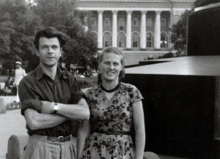 Melnbalta fotogrāfija ar jaunu vīrieti un sievieti, kas stāv parkā pilsētvidē; fonā redzama monumentāla ēka ar kolonnām.
