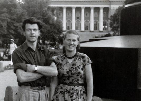 Melnbalta fotogrāfija ar jaunu vīrieti un sievieti, kas stāv parkā pilsētvidē; fonā redzama monumentāla ēka ar kolonnām.