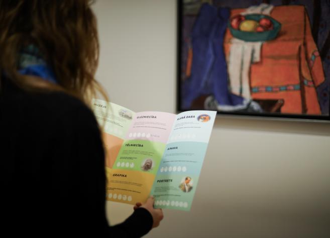 Cilvēks rokās tur vairākas krāsainas drukātas kartītes ar uzrakstu “Sveiks muzejā!”.