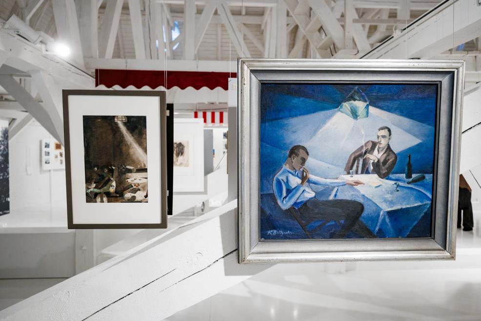 Exhibition space with suspended artworks; in the foreground, a blue-toned figurative painting depicting two people seated at a table.
