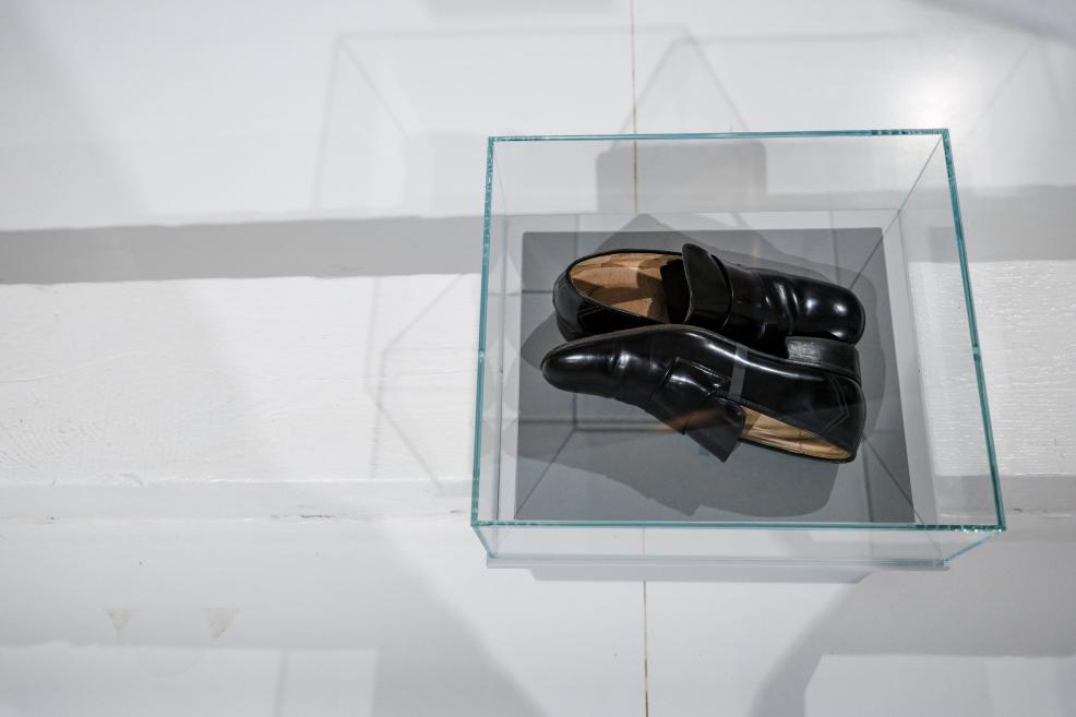 Art object displayed in a glass case – black leather shoes placed on a grey base in an exhibition space.
