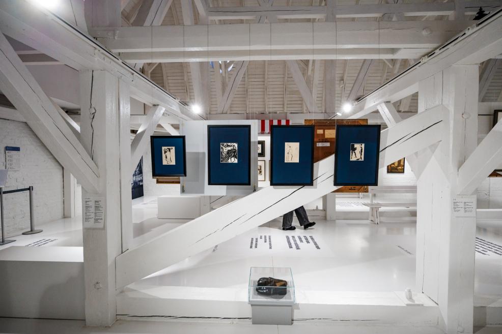 Exhibition view in a bright gallery space with white wooden beams, framed graphic works in blue mounts and an art object displayed in a glass case.