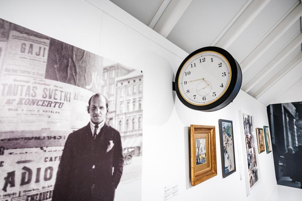 Exhibition display detail featuring a wall clock, a historical black-and-white photograph on the left, and several framed artworks on a white wall.