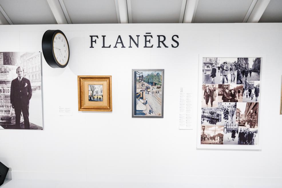 Exhibition display section titled “Flâneur”, featuring a wall clock, paintings, and a collage of historical photographs on a white wall.