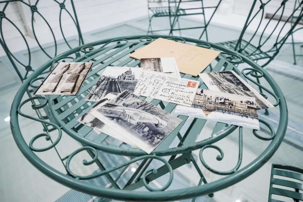 Detail of a café-themed exhibition display showing a round metal table with vintage postcards and an envelope, set against a glass floor.