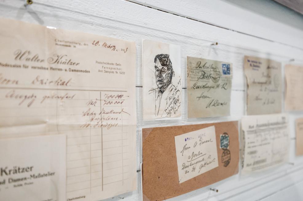 Museum exhibition wall displaying early 20th-century documents, letters, envelopes, and cards with handwritten notes, stamps, and a small graphic portrait sketch.