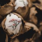 Tuvplānā redzami brūni čaulu veidojumi ar ievietotiem auduma fragmentiem, uz kuriem izšūti smalki augu motīvi.