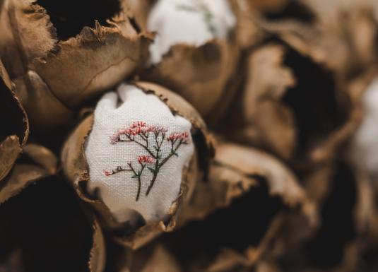 Tuvplānā redzami brūni čaulu veidojumi ar ievietotiem auduma fragmentiem, uz kuriem izšūti smalki augu motīvi.