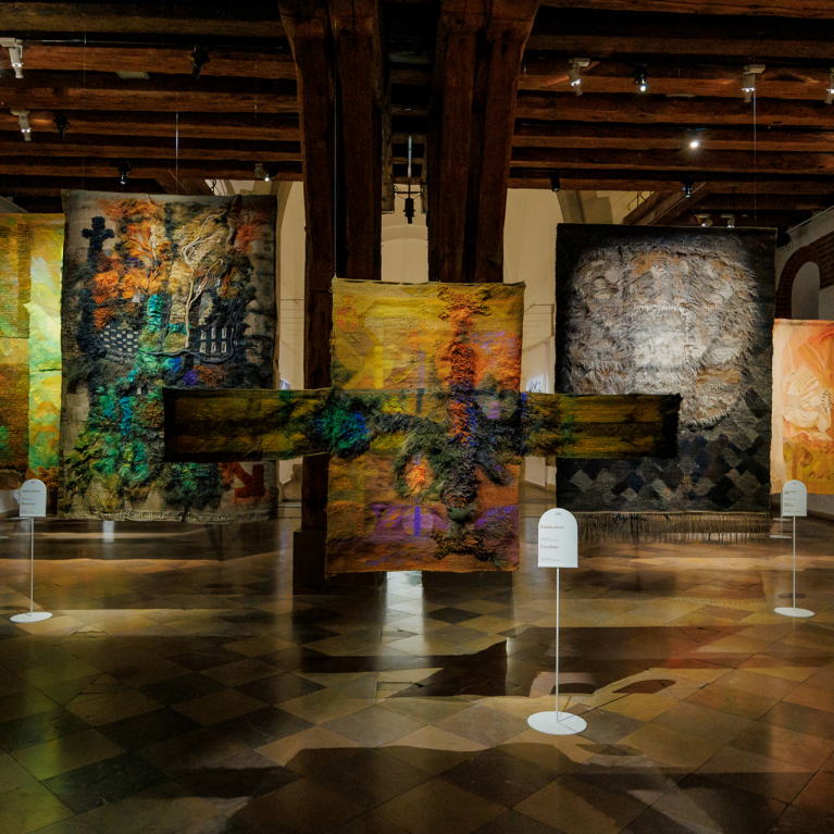 An exhibition hall featuring large-scale textile artworks in various colours and textures, suspended from the ceiling between wooden beams.