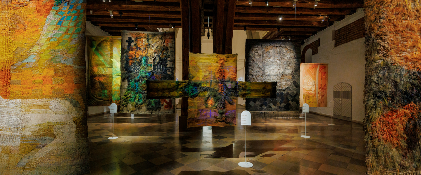 An exhibition hall featuring large-scale textile artworks in various colours and textures, suspended from the ceiling between wooden beams.