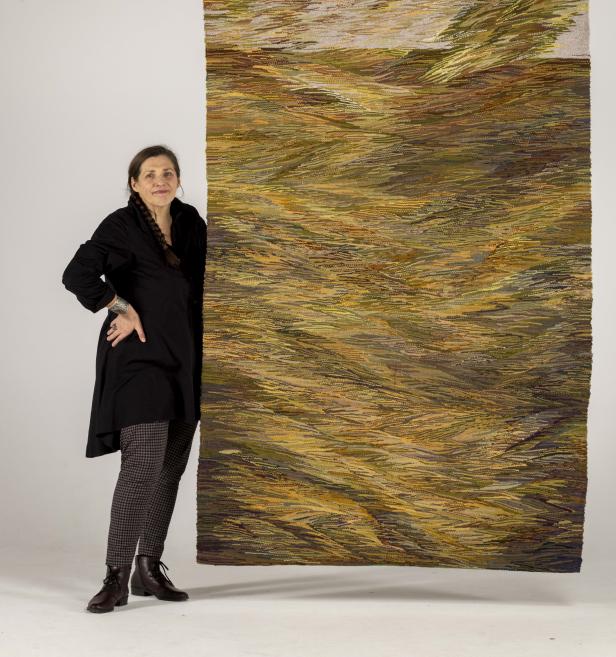 "Artist standing beside a large woven tapestry with flowing earth-toned patterns resembling a landscape.