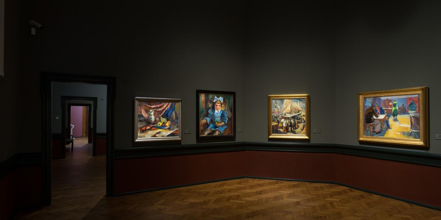 Muzeja zālē pie sienas izstādītas vairākas gleznas ar apgaismojumu.