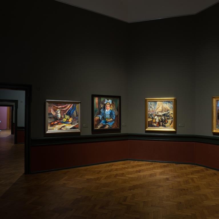 Muzeja zālē pie sienas izstādītas vairākas gleznas ar apgaismojumu.