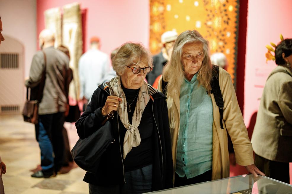 Two elderly women view a glass display case in a museum exhibition, with other visitors and brightly colored pink walls with artworks in the background.