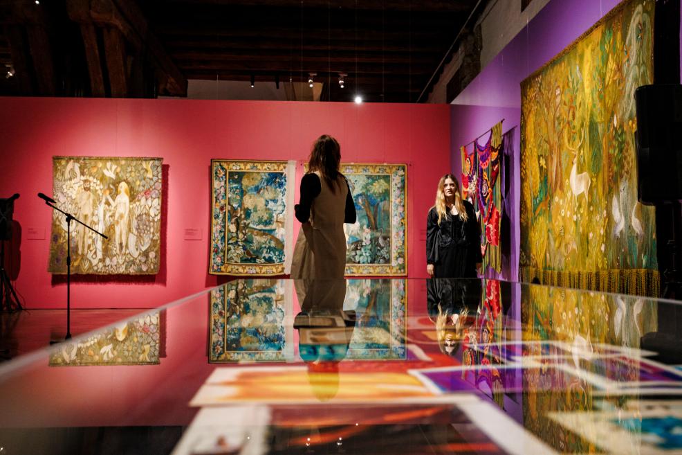 Colorful textile works hang in a museum gallery with pink and purple walls, with two women standing near a reflective table in the foreground.