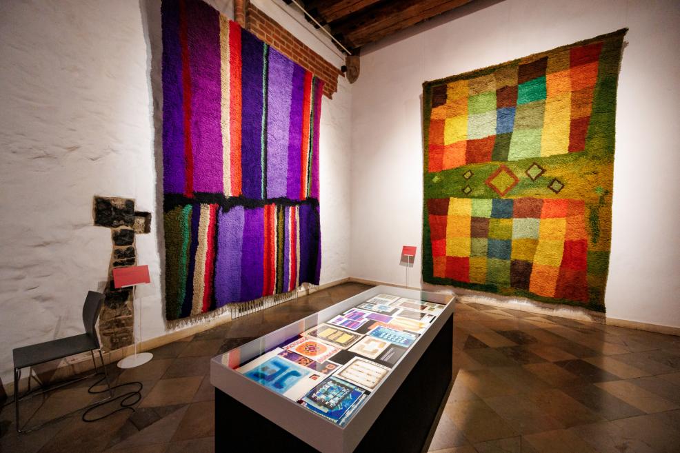 In a museum gallery, two large colorful textile wall hangings and a glass display case with objects on a table are visible.