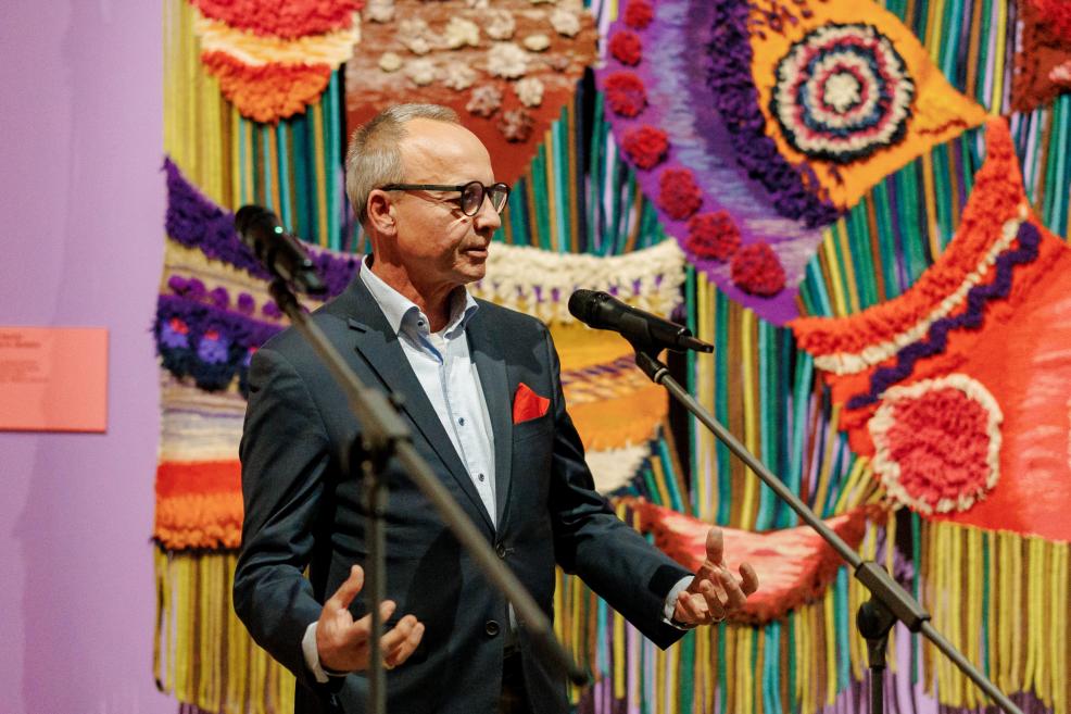 A man in a suit and glasses speaks at a microphone in front of a colorful textile mosaic.