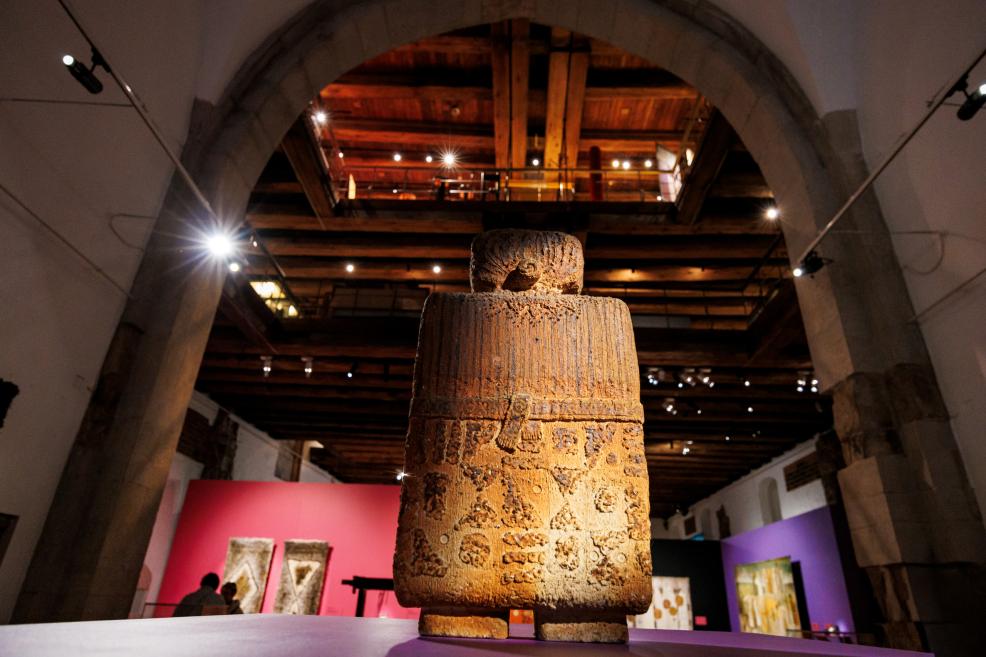 A small cylindrical wooden sculpture with a relief-decorated surface stands on a pedestal in a museum hall beneath a large arch, lit by spotlights.