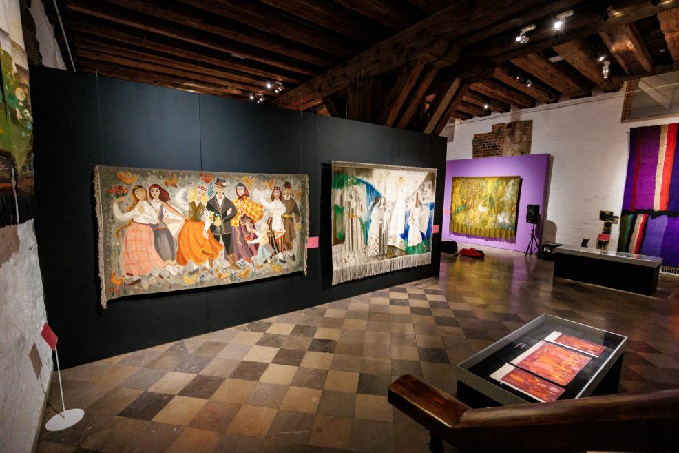 Museum exhibition hall with several colorful tapestries displayed on the walls, visible wooden beams, a tiled floor, and a display case with works in the foreground.
